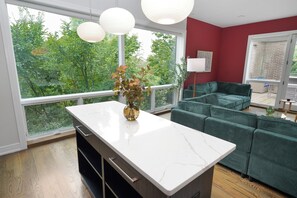 A modern living room with a large window, a green sofa set, and a marble kitchen island. - Little Italy 4BR Retreat w/ Optional Parking (Chicago)