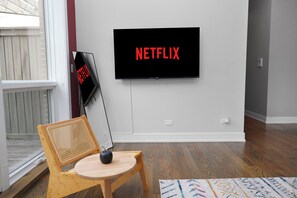A room with a television mounted on the wall displaying 'NETFLIX' in red letters, a wooden chair with a woven backrest, and a small round table with an apple on it.