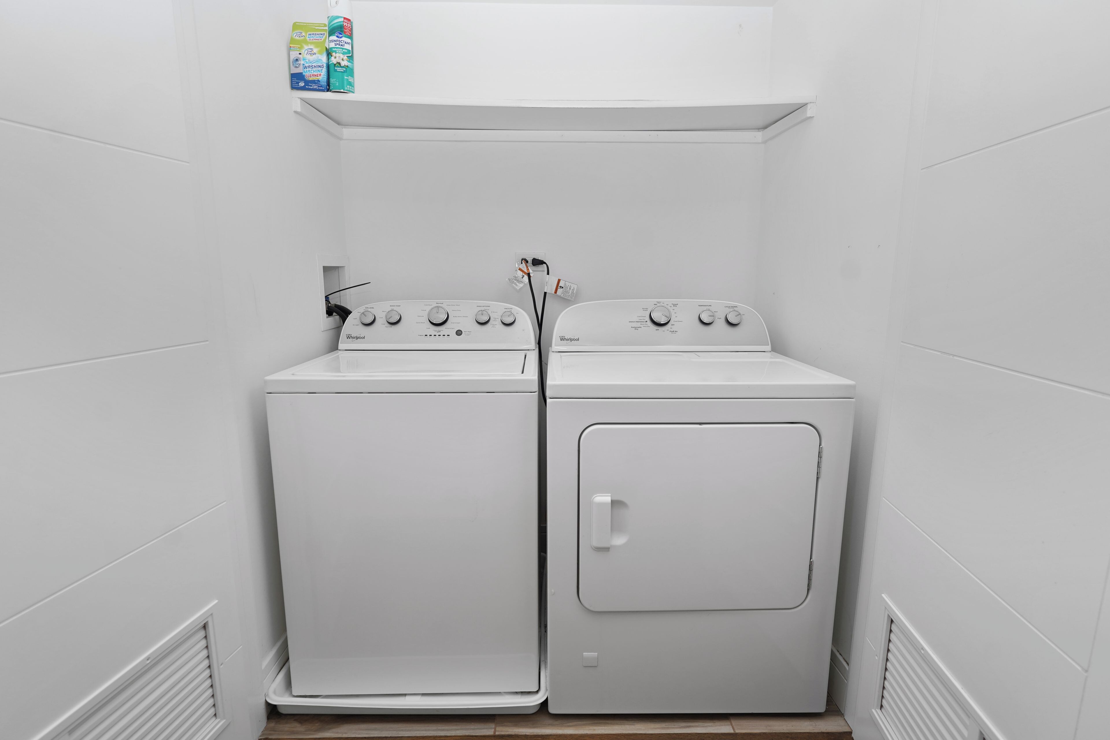 A laundry room with a front-loading washer and dryer, a shelf with detergent bottles, and a vent on the wall.