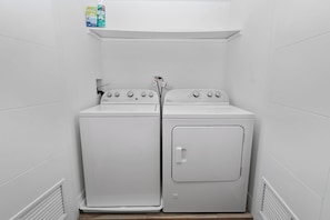 A laundry room with a front-loading washer and dryer, a shelf with detergent bottles, and a vent on the wall. - Little Italy 4BR Retreat w/ Optional Parking (Chicago)