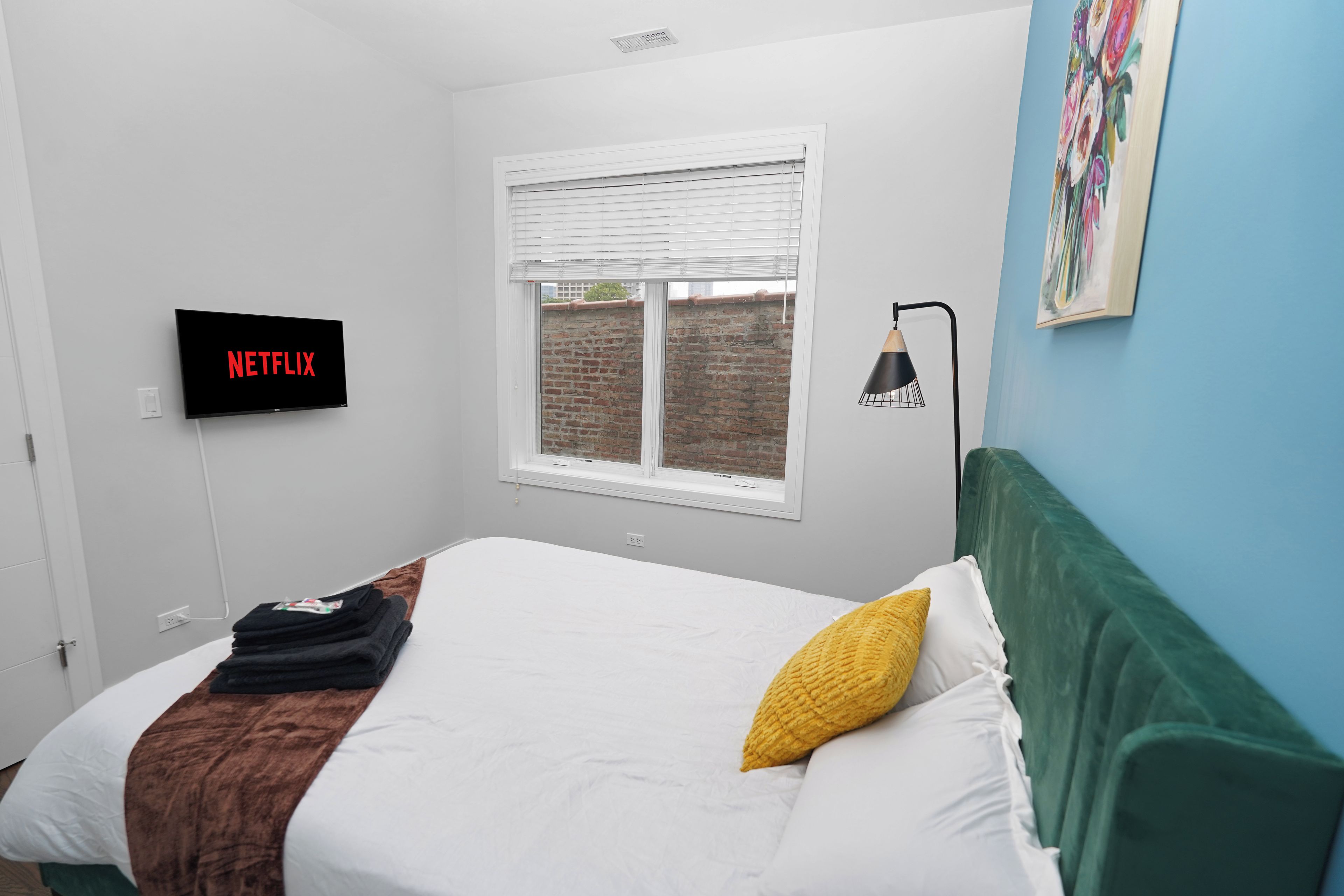 A bedroom with a bed, a wall-mounted TV displaying 'NETFLIX', a window with blinds, and a painting on the wall.
