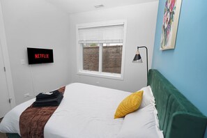 A bedroom with a bed, a wall-mounted TV displaying 'NETFLIX', a window with blinds, and a painting on the wall. - Little Italy 4BR Retreat w/ Optional Parking (Chicago)
