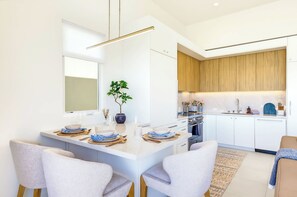 A modern kitchen with white cabinetry, a central island, and a dining area with chairs and a table set for two. - Sunset House - Rooftop Ocean & Bay Views (San Diego)