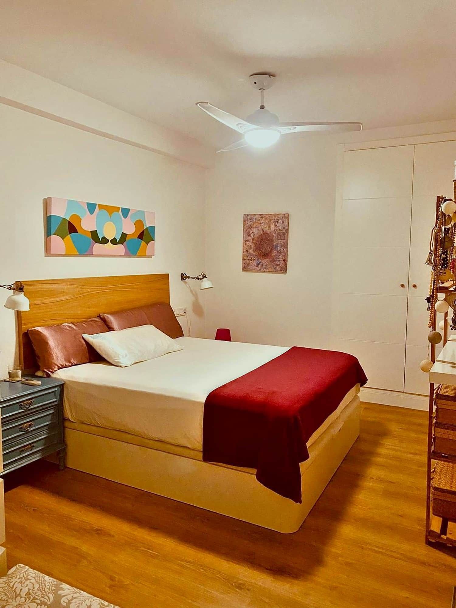 A bedroom with a bed, a nightstand, a lamp, a ceiling fan, and artwork on the walls.