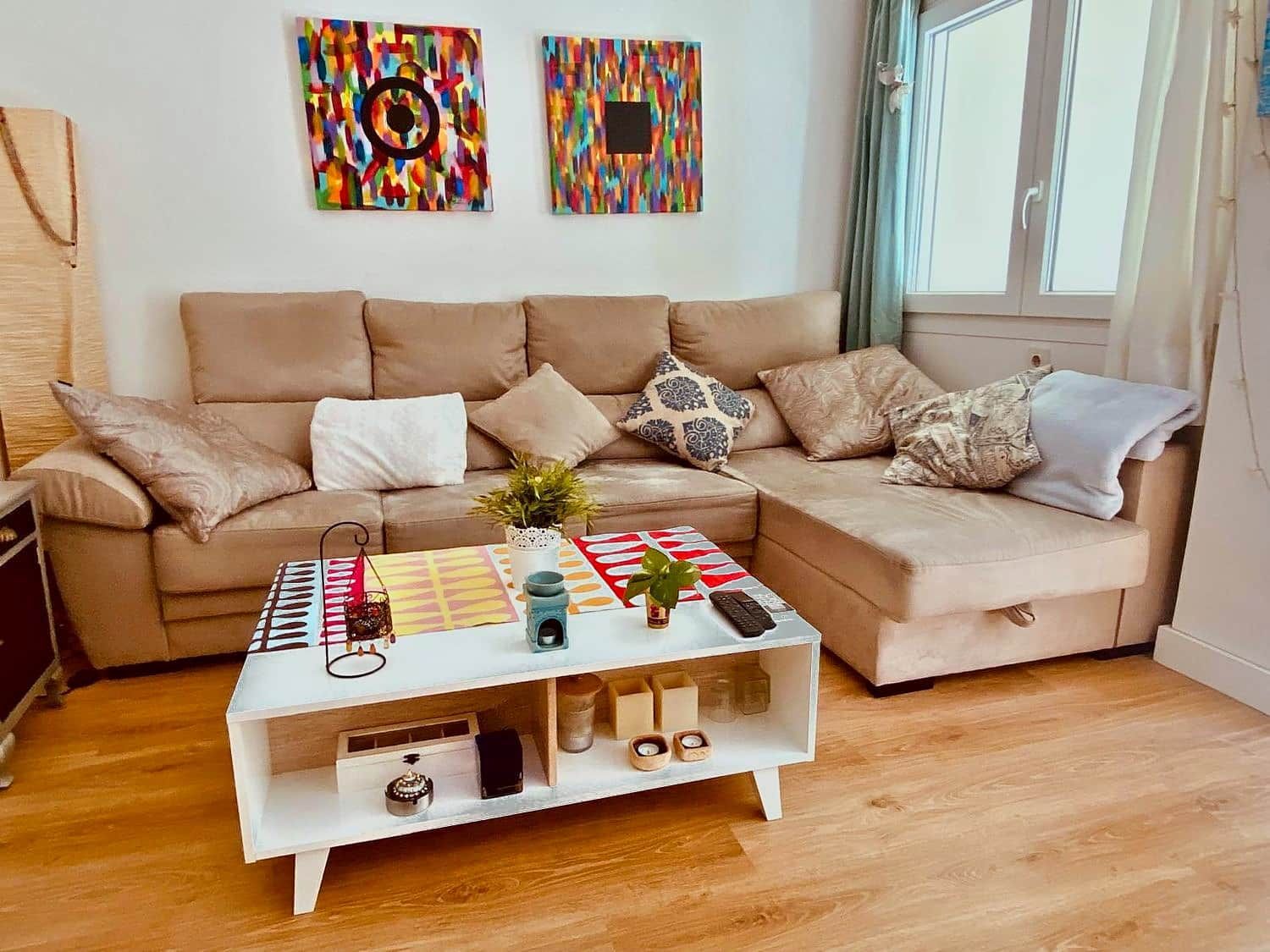 Una sala de estar moderna con un sofá color beige, una mesa de centro blanca con un patrón geométrico colorido y dos cuadros abstractos en la pared.