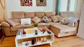 Una sala de estar moderna con un sofá color beige, una mesa de centro blanca con un patrón geométrico colorido y dos cuadros abstractos en la pared.