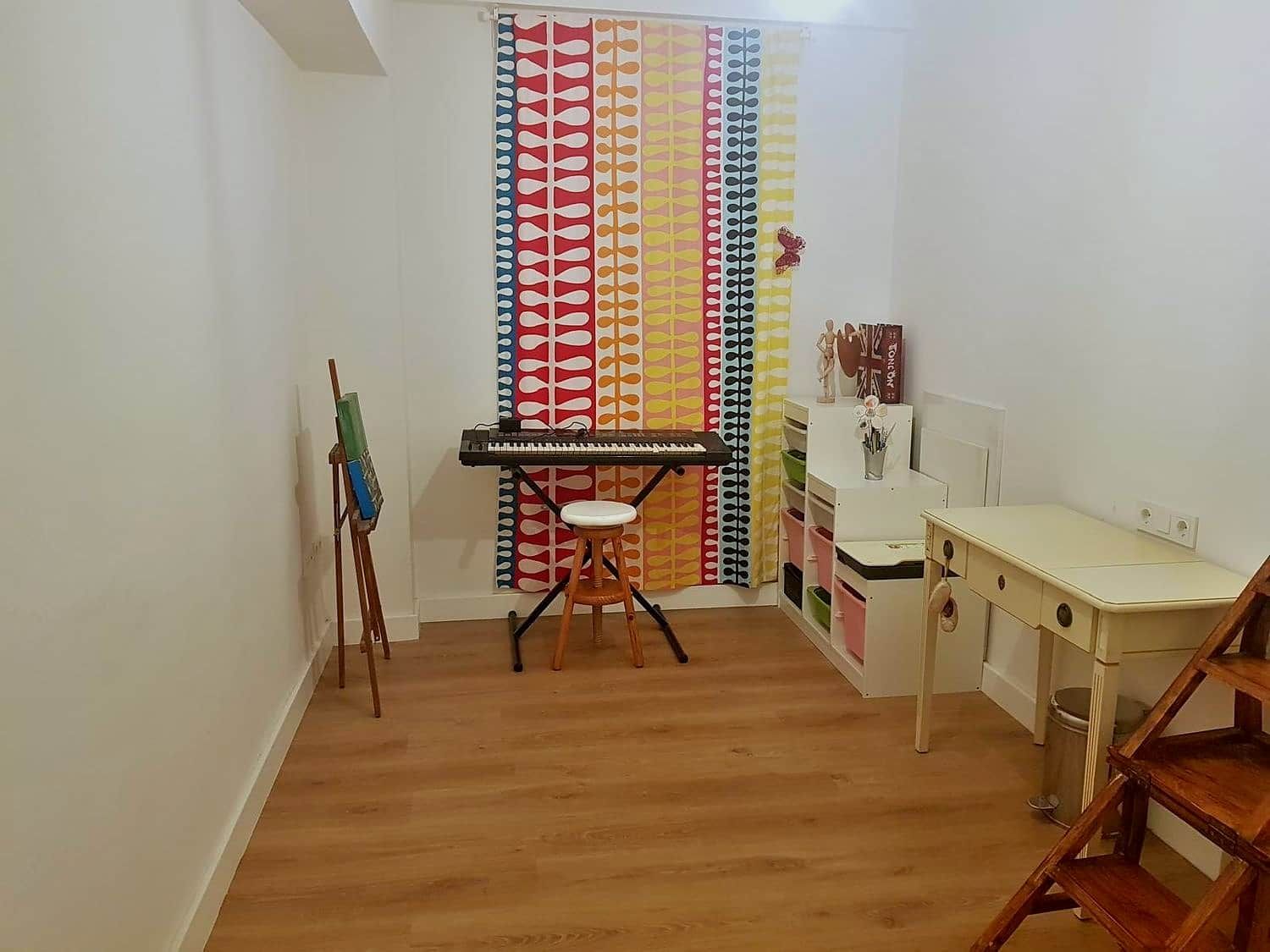 A room with a wooden floor, a white desk, a stool, a keyboard, a painting on an easel, and a colorful curtain.