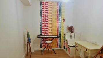 A room with a wooden floor, a white desk, a stool, a keyboard, a painting on an easel, and a colorful curtain.