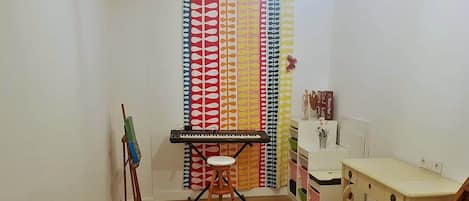 A room with a wooden floor, a white desk, a stool, a keyboard, a painting on an easel, and a colorful curtain.