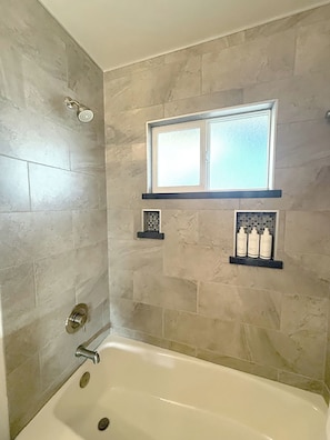 A bathroom with a bathtub, a window, and a shelf with toiletries. - Presidential Two Bedroom, Two Bath Suite (San Luis Obispo)