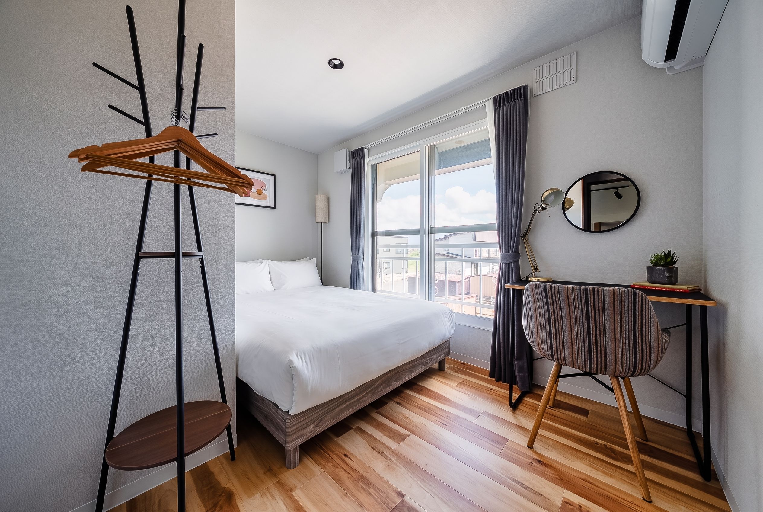 A hotel room with a bed, a desk with a chair, a hanging coat rack, a window with curtains, and a ceiling light.