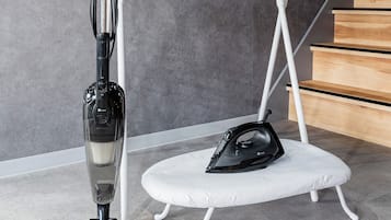 Una habitación con una aspiradora, una tabla de planchar y un estante metálico blanco junto a una pared gris.