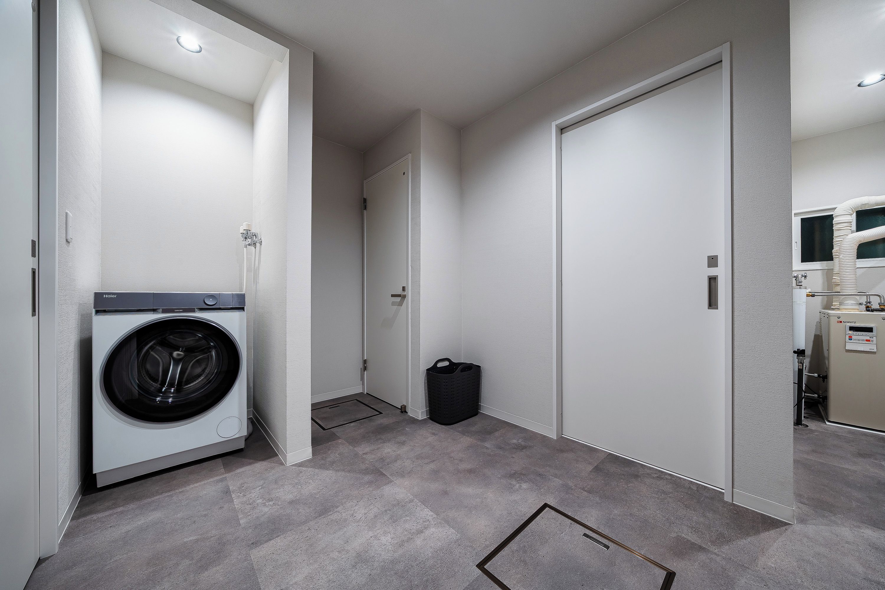 A laundry room with a front-loading washer, a trash bin, and a door leading to another room.