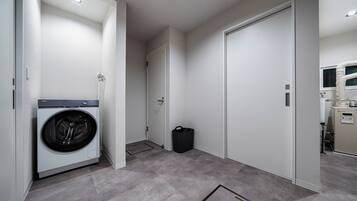 A laundry room with a front-loading washer, a trash bin, and a door leading to another room.