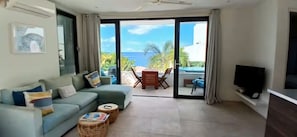 A living room with a sofa, a television, and a view of the ocean through a sliding glass door. - Indigo Sweet is a highly rated 1-bedroom apartment located in Indigo Bay. (Cole Bay)