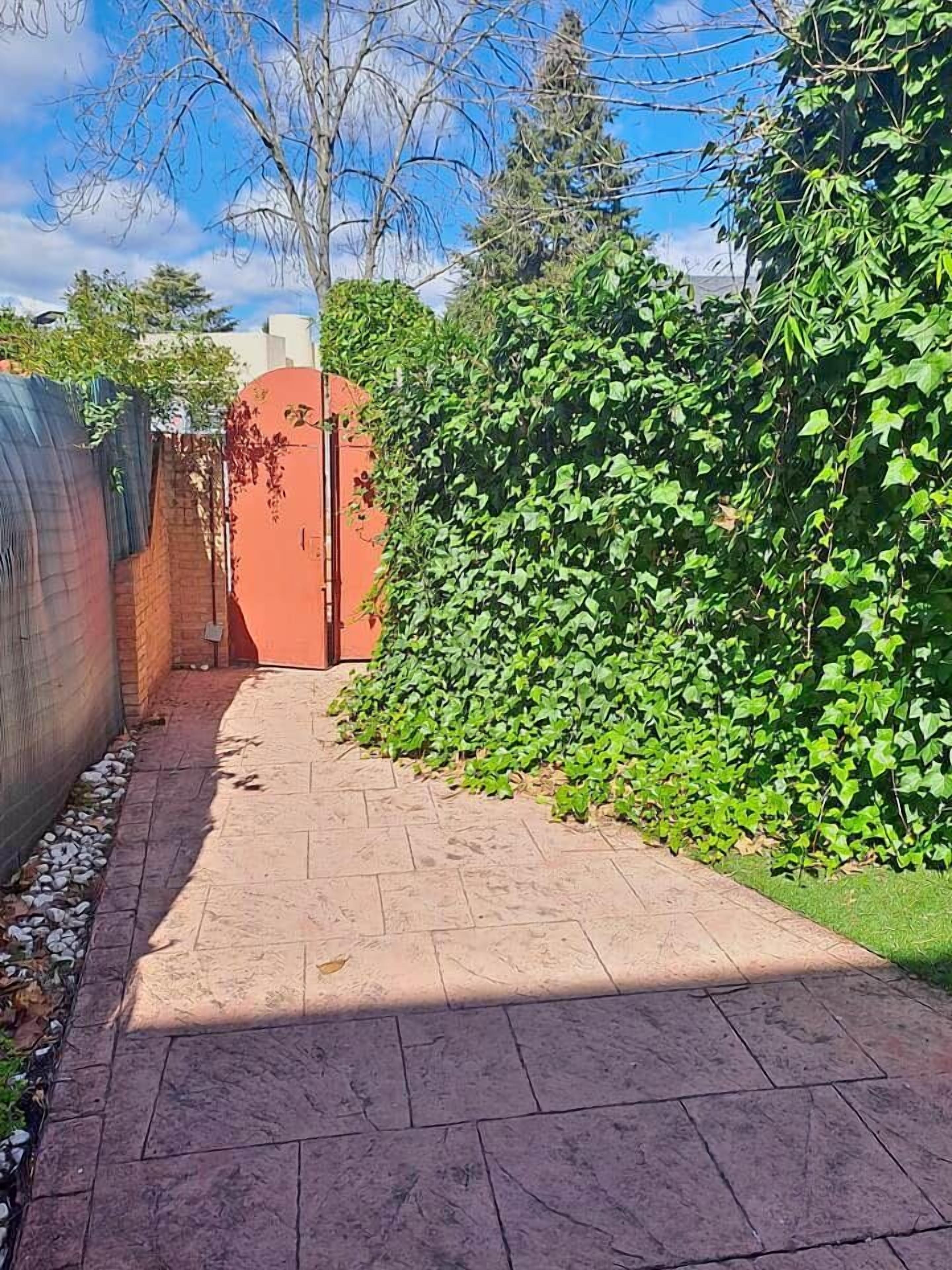 Un sendero pavimentado que lleva a una reja roja, rodeado de abundante follaje verde y un muro de ladrillo.
