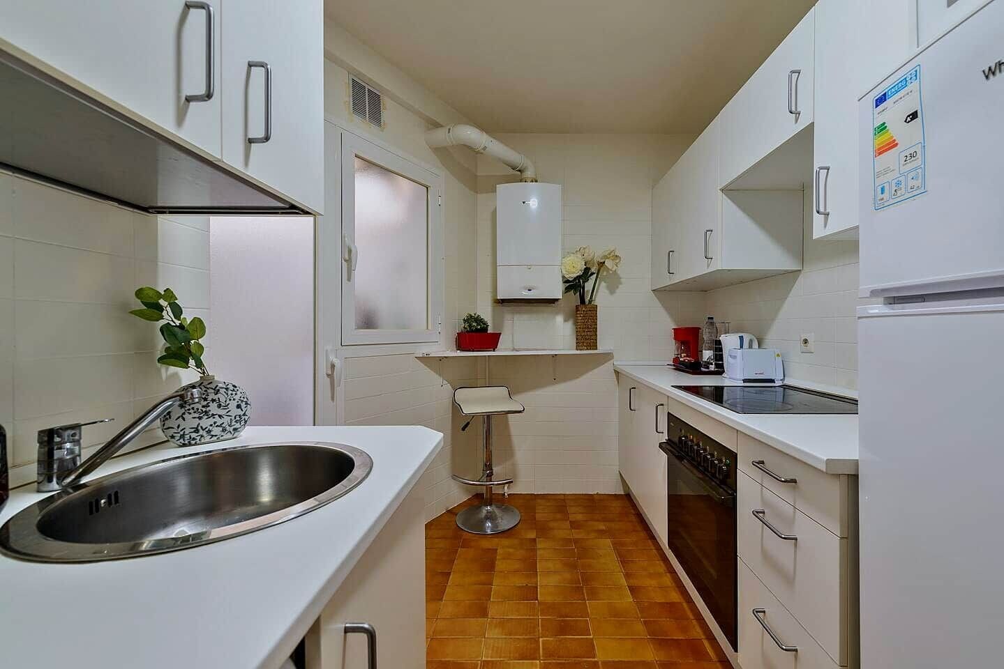 A compact kitchen with white cabinets, a stainless steel sink, a small stool, and a tiled floor.