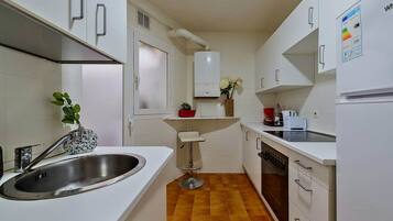 A compact kitchen with white cabinets, a stainless steel sink, a small stool, and a tiled floor.