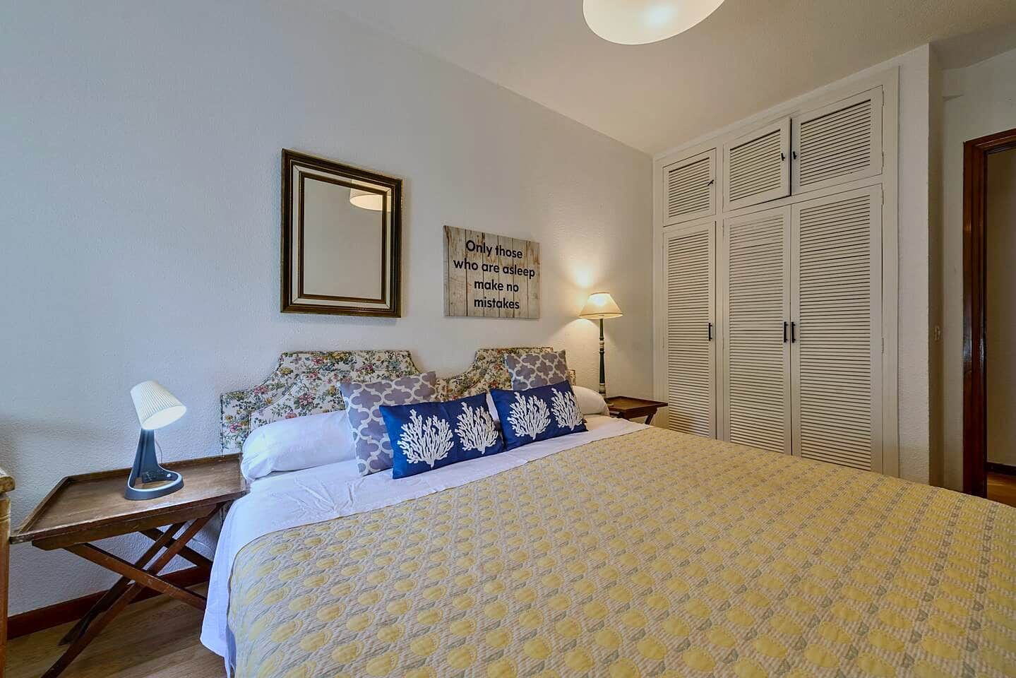 Un dormitorio con cama, lámpara de noche, mesita de noche, espejo montado en la pared y armario con puertas batientes.