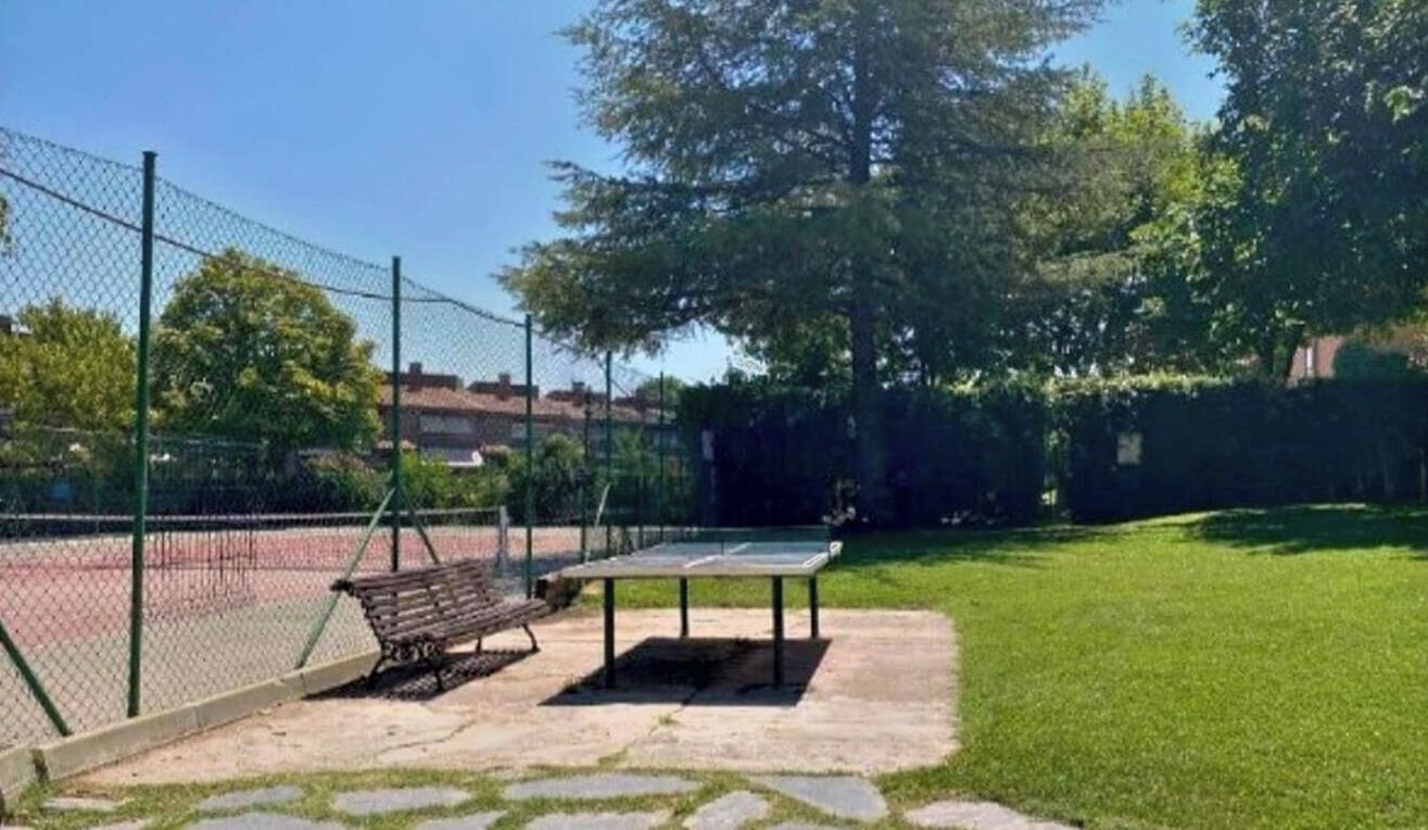 Cancha de tenis con un banco y una mesa, rodeada por una cerca de malla y follaje.