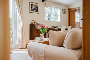 A cozy living room with a sofa, a wooden cabinet, a table with a vase of flowers, and a window with blinds. - Hoglets - Cosy Countryside Retreat (Conderton)