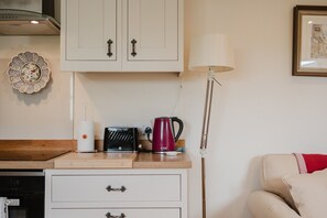 A kitchenette with a toaster, kettle, and a lamp. There is a cabinet above the counter and a framed picture on the wall. - Hoglets - Cosy Countryside Retreat (Conderton)