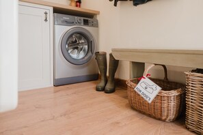 A laundry area with a washing machine, a pair of boots, and a basket labeled 'For Dog's'. - Hoglets - Cosy Countryside Retreat (Conderton)