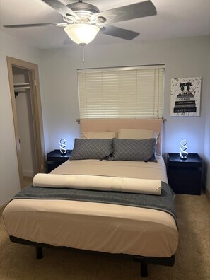 Une chambre à coucher avec un lit, un ventilateur de plafond, une fenêtre avec des stores et un mur orné d’une affiche.