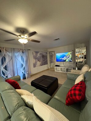 A living room with a green sofa, a black ottoman, a ceiling fan, and a television displaying a scenic landscape. - Maison Grape  Elegant 3BR Home near Medical Center & NRG (Houston)
