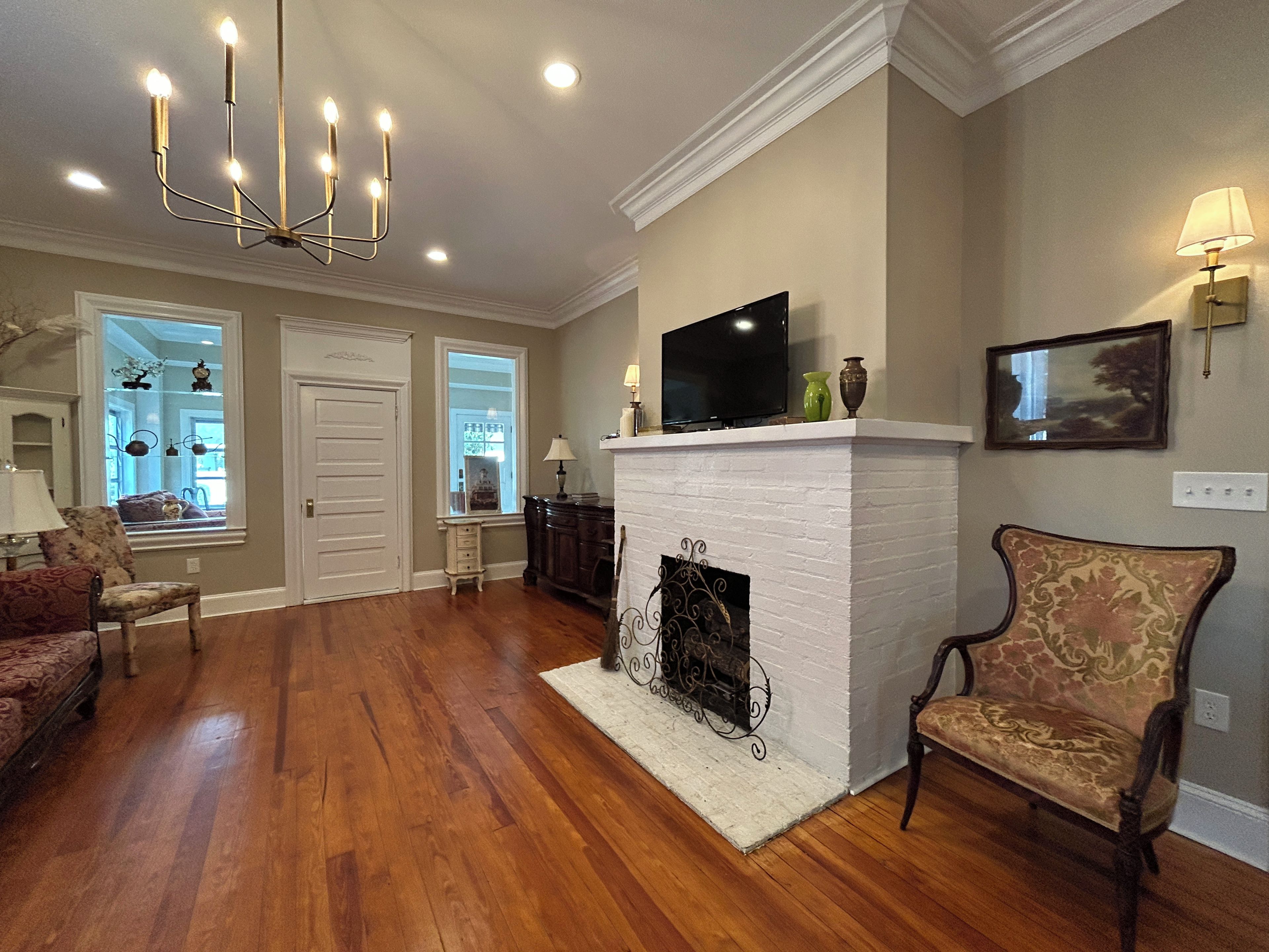 A living room with hardwood flooring, a white fireplace, a television on the mantel, and a chandelier.