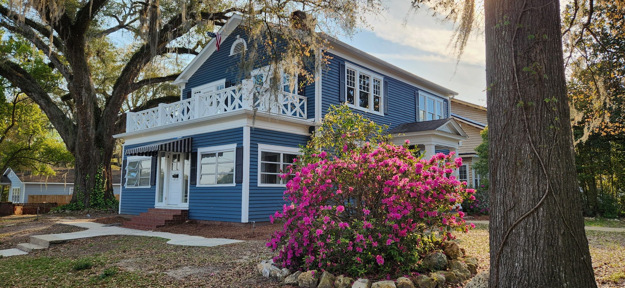 A two-story house with a porch, surrounded by trees and a flowering bush.