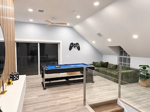 A modern living room with a pool table, a ceiling fan, and a gaming console on the wall.