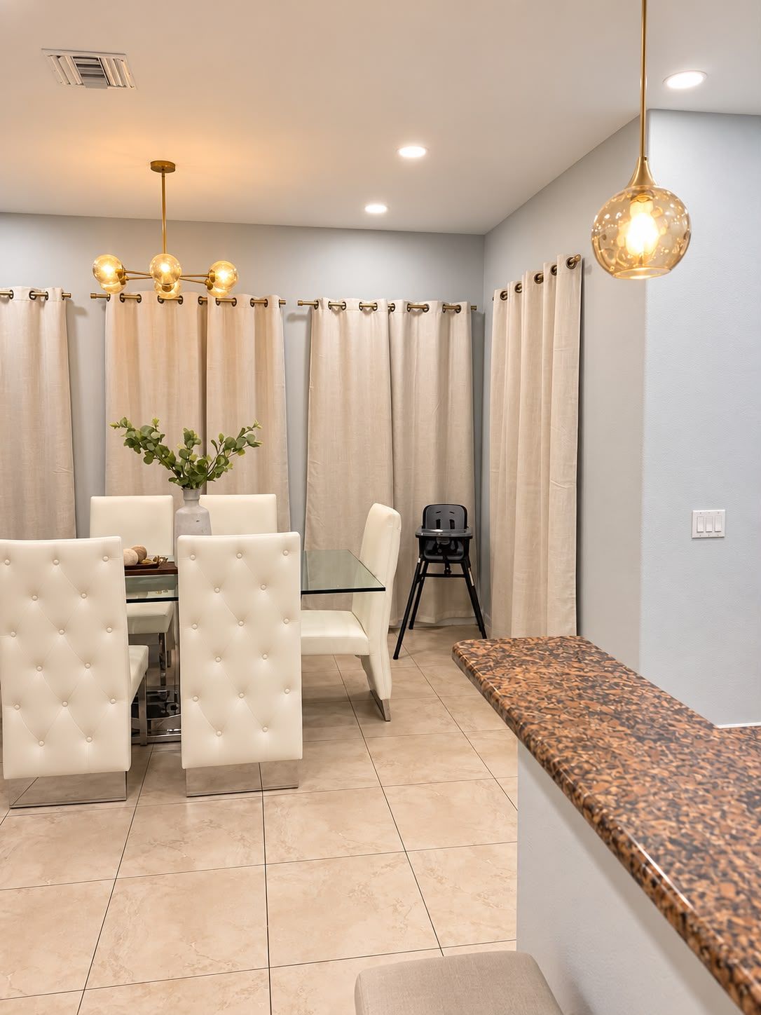 A dining area with a glass table, white upholstered chairs, a black folding chair, and a granite countertop.