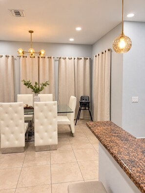 A dining area with a glass table, white upholstered chairs, a black folding chair, and a granite countertop.
