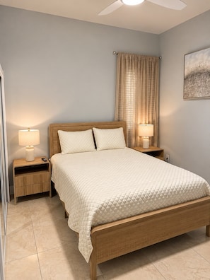 A bedroom with a bed, two bedside tables with lamps, a ceiling fan, a window with curtains, and a wall-mounted artwork.