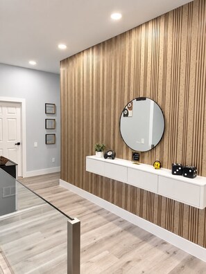 Un baño moderno con pared de paneles de madera, un espejo redondo, un tocador blanco y una cabina de ducha de vidrio.