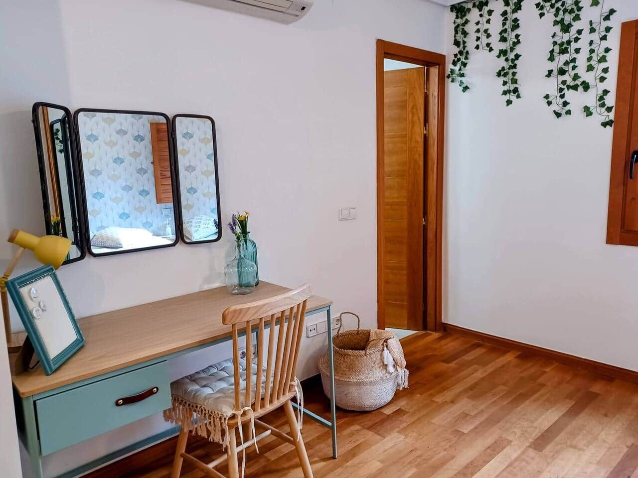Een kamer met een houten bureau, een stoel, een klein tafeltje en een spiegel aan de muur. De deur heeft een houten omlijsting en er staat een mand op de vloer.