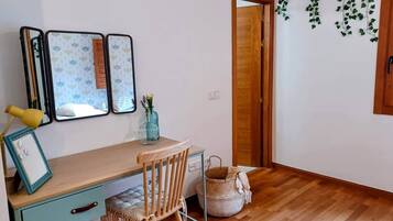 Een kamer met een houten bureau, een stoel, een klein tafeltje en een spiegel aan de muur. De deur heeft een houten omlijsting en er staat een mand op de vloer.