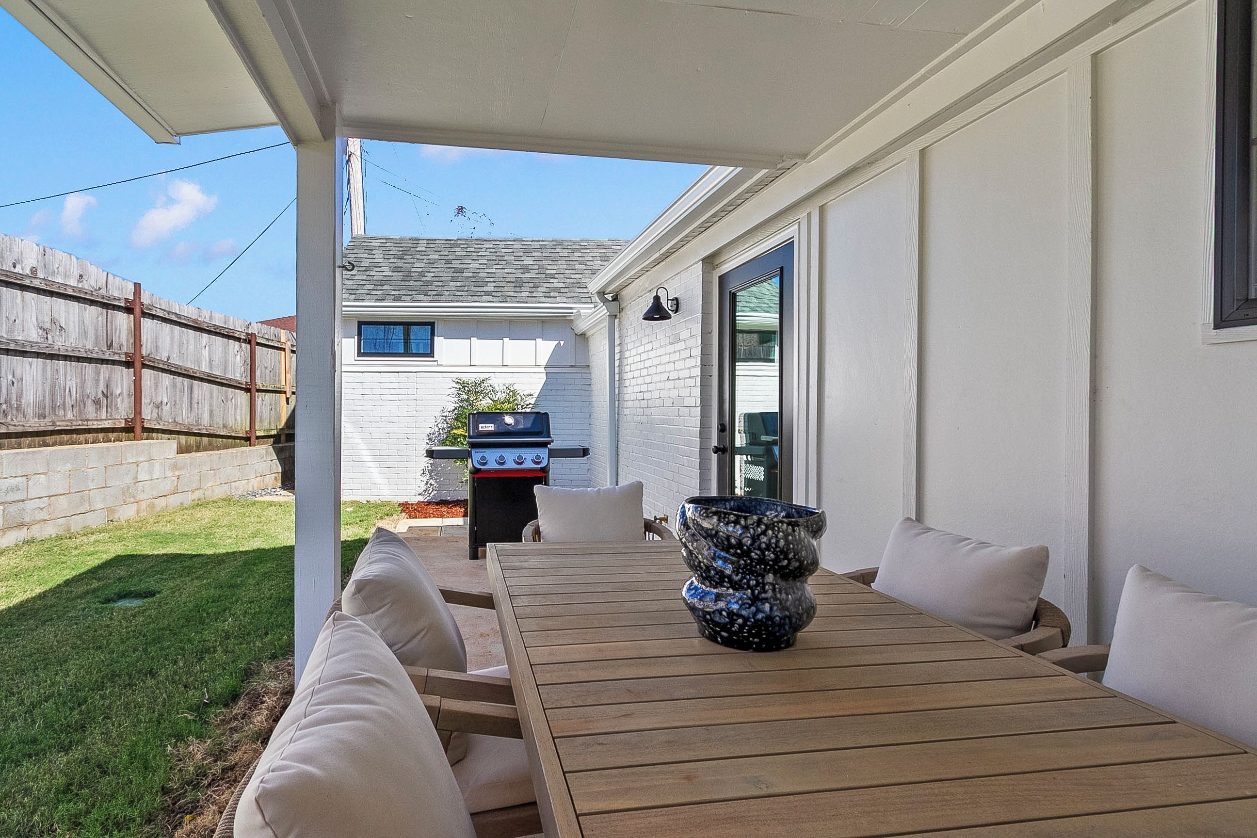 Een terras buiten met een houten tafel en witte kussens, een barbecue en een decoratieve vaas.