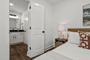Et hotelværelse med seng, et natbord, en lampe, et maleri, et badeværelse med et spejl og en vask.