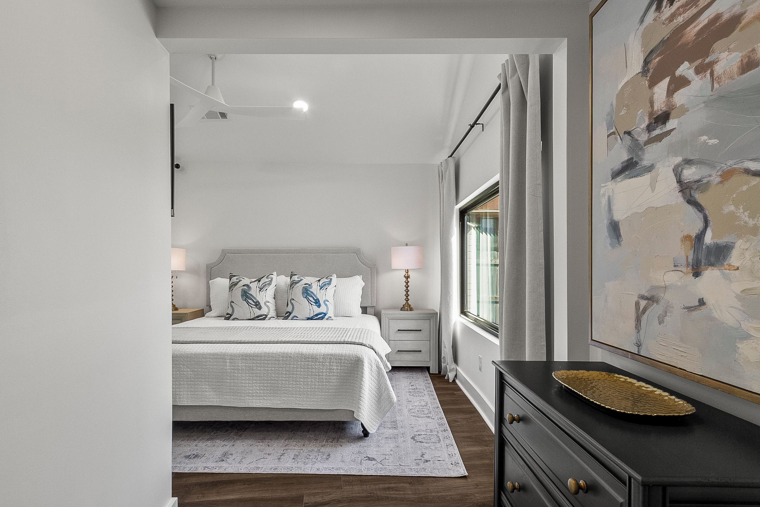 A bedroom with a bed, bedside tables, a dresser, a window with curtains, and a painting on the wall.