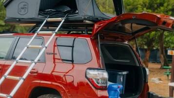 Um SUV vermelho com uma tenda no teto, uma cadeira dobrável branca, um cooler vermelho e uma mesa com equipamentos de camping.