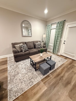 Un salon comprenant un canapé, une table basse et des poufs, un miroir au mur et une porte avec des rideaux.