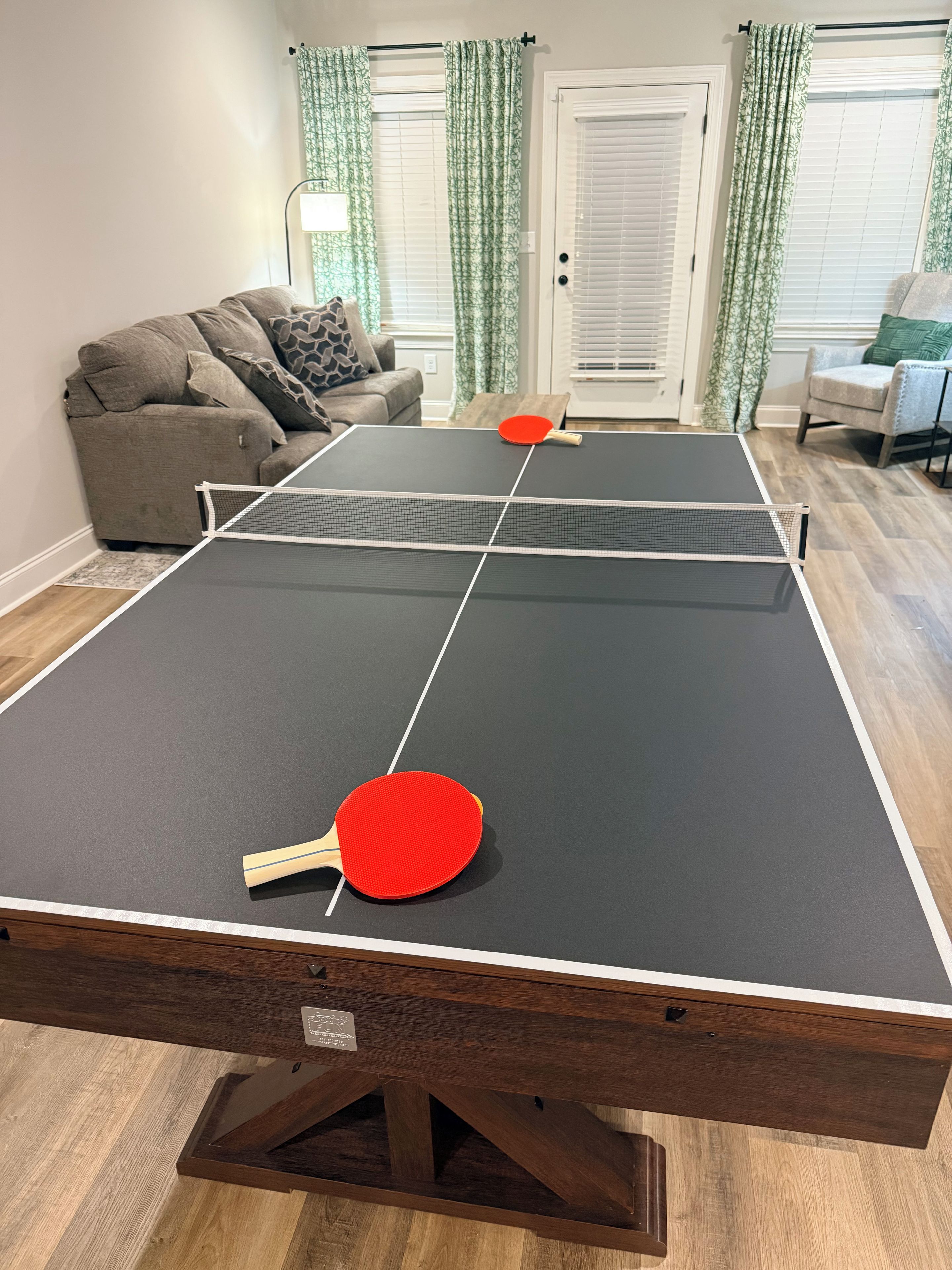 Una mesa de ping pong con dos palas, un sofá y una silla en una habitación con cortinas estampadas y una puerta.