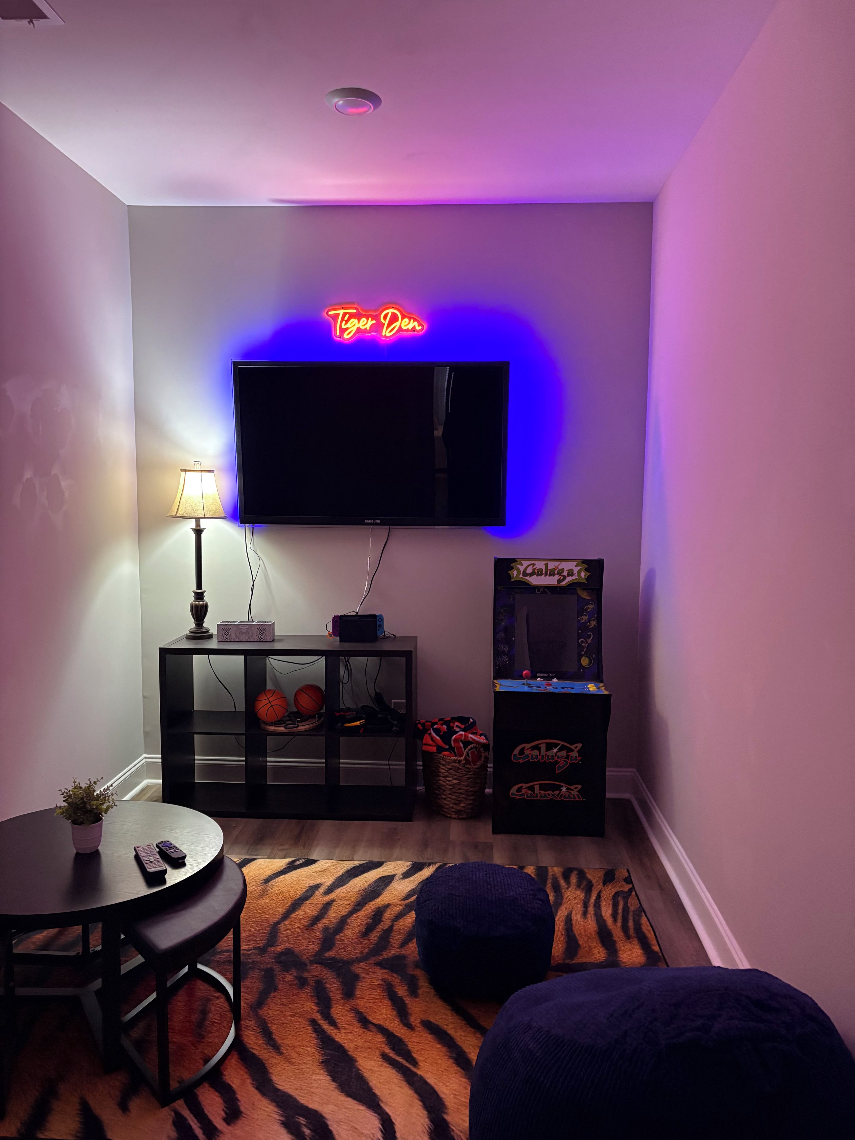 A living room with a flat-screen TV, a neon sign, a lamp, a gaming console, a basketball, and a tiger-patterned rug.