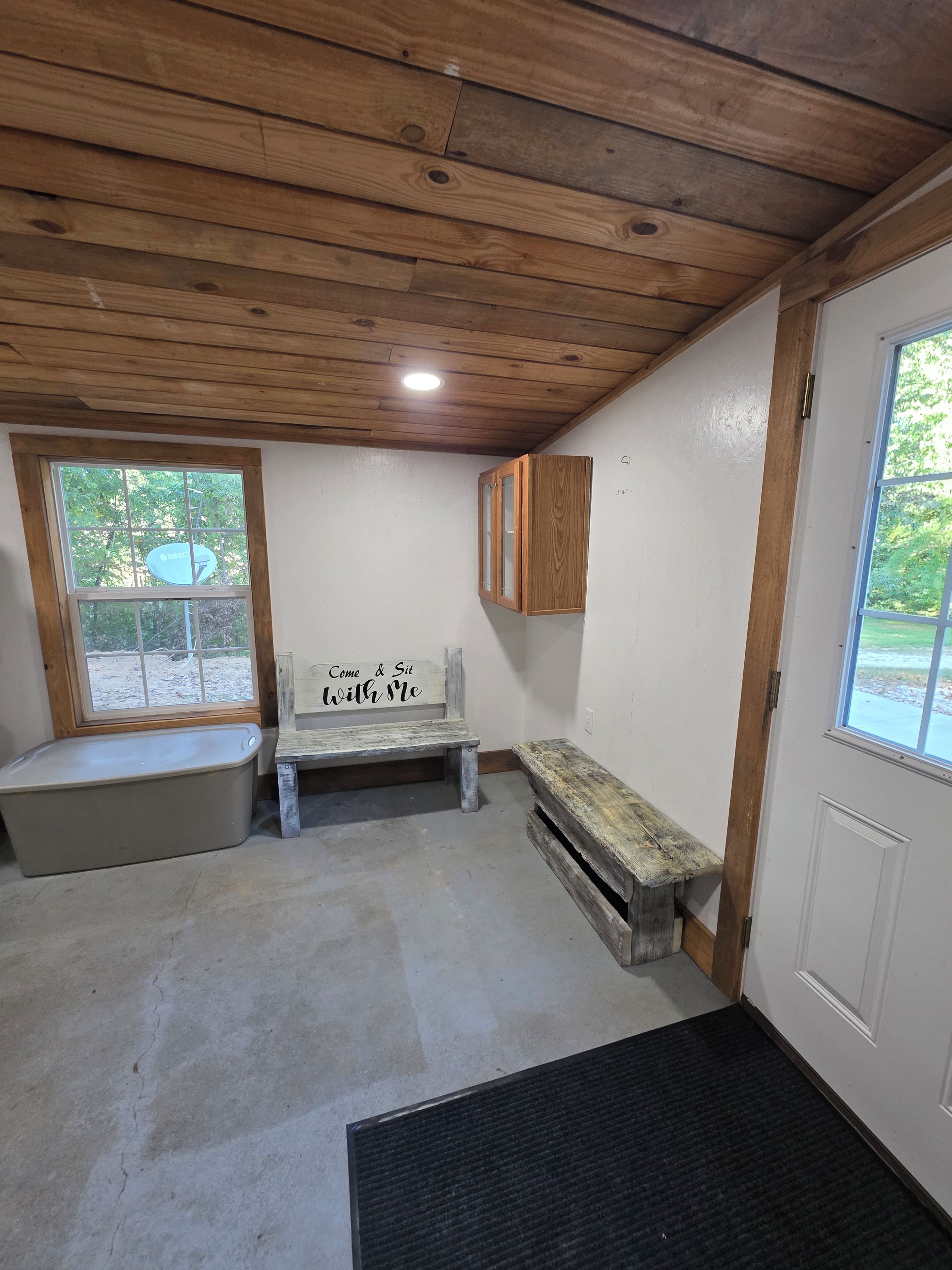A room with a wooden ceiling, a bench, a cabinet, a bench with a sign, and a door.