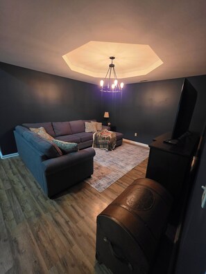 A modern living room with a sectional sofa, a small coffee table, a leather ottoman, a television, and a unique ceiling light fixture. - Quiet 3-Bedroom Country Stay in Nebo, Illinois (Nebo)