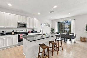 A modern kitchen with white cabinetry, a central island with stools, a dining area with a glass table, and a view through a large window. - Best location near Orlando’s world famous attractions (Kissimmee)