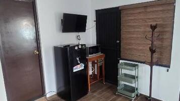 Quarto com piso de madeira, porta fechada, armário preto, televisão fixada na parede e janela com persiana.