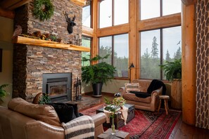 A cozy living room with a fireplace, leather furniture, and large windows offering a view of trees.